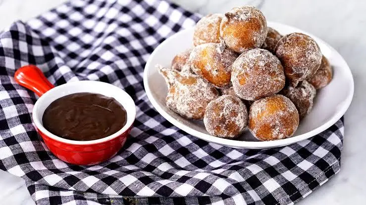 Um prato branco com bolinhos de chuva em cima e ao lado um potinho vermelho com brigadeiro, embaixo dos dois um pano xadrez preto e branco