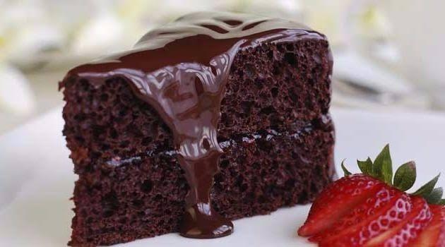 Um pedaço de bolo de chocolate com a calda caindo, em um prato branco e um morango ao lado para enfeite