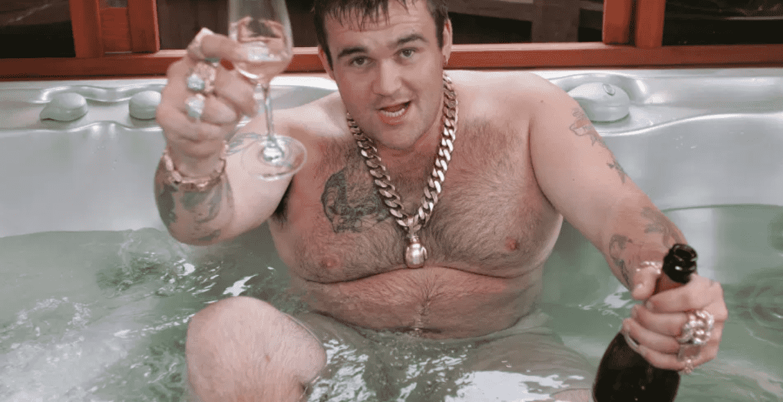 Foto mostra homem adulto branco, sem camisa sentando em uma banheira, com uma taça na mão direita e uma garrafa de espumante na esquerda. Ele usa colares e anéis dourados.
