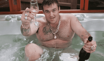 Foto mostra homem adulto branco, sem camisa sentando em uma banheira, com uma taça na mão direita e uma garrafa de espumante na esquerda. Ele usa colares e anéis dourados.