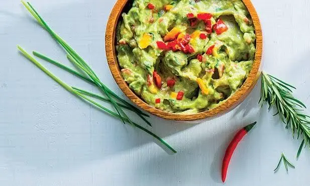 Foto de cima, mostrando um guacamole em um recipiente de madeira clara e uma pimenta na mesa