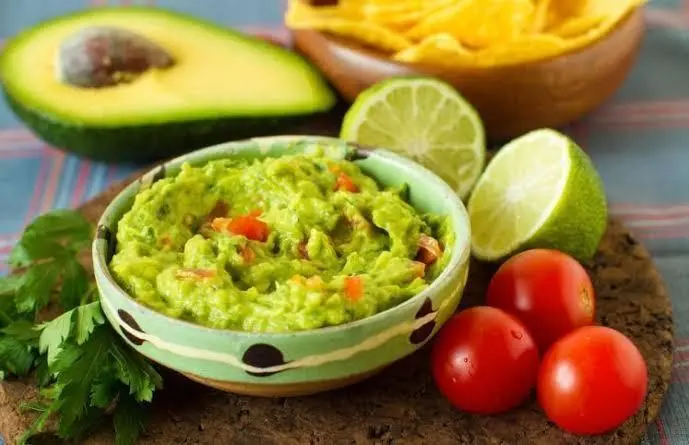 Guacamole em um pote de porcelana com bolinhas e em volta três tomates e um limão cortado ao meio