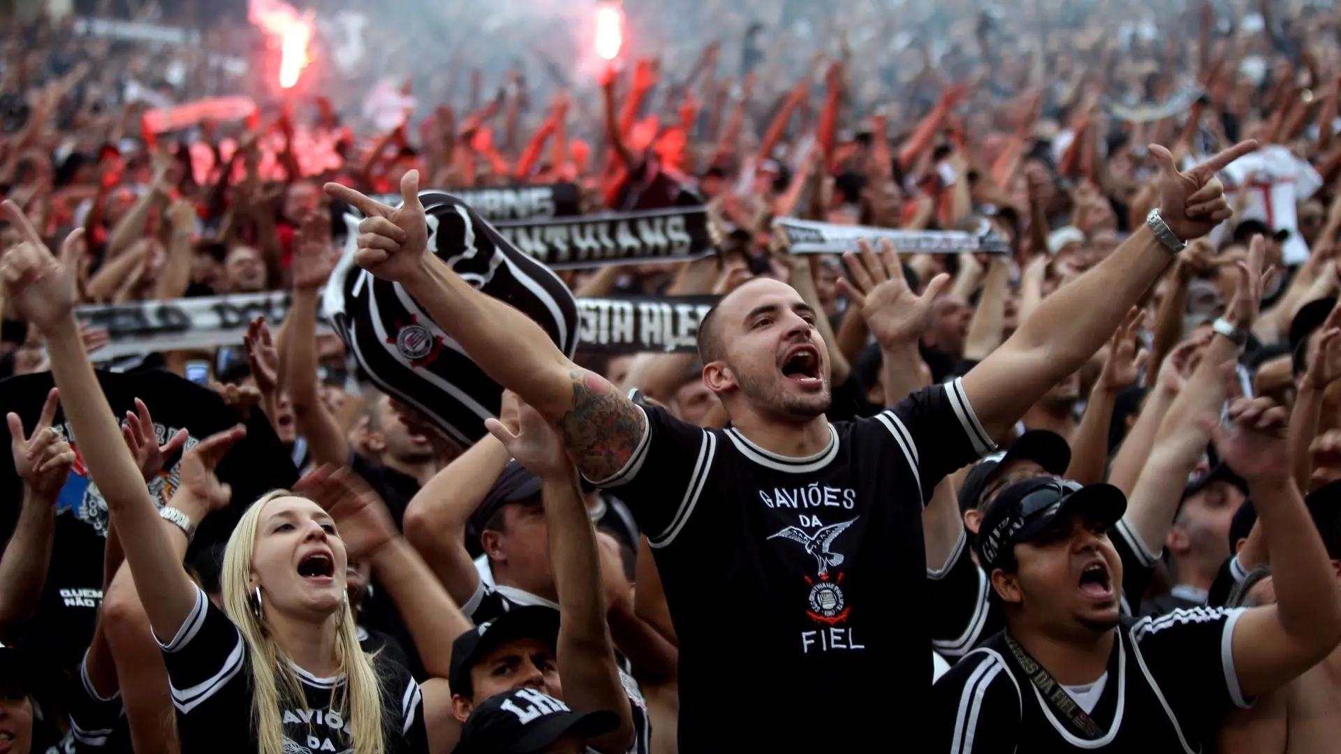 Torcida do Corinthians cantando, na frente um homem branco esticando os punhos 