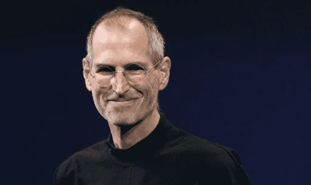 Foto de Steve Jobs, homem adulto, de cabelos grisalhos, usando óculos e uma roupa preta