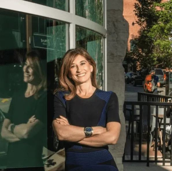 Foto mostra mulher adulta usando vestido preto, encosta em uma porta de vidro.