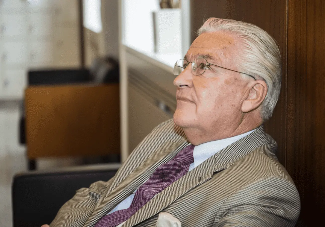 Foto do ex-milionário sentado de perfil. Homem branco, vestindo terno e usando óculos, de cabelos brancos.