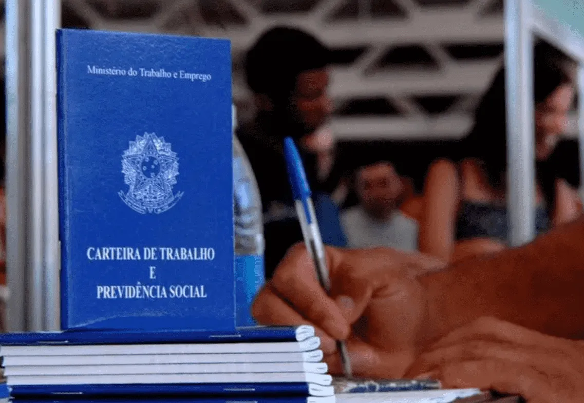 Imagem mostra uma carteira de trabalho em pé e uma mãe assinando um papel a caneta.