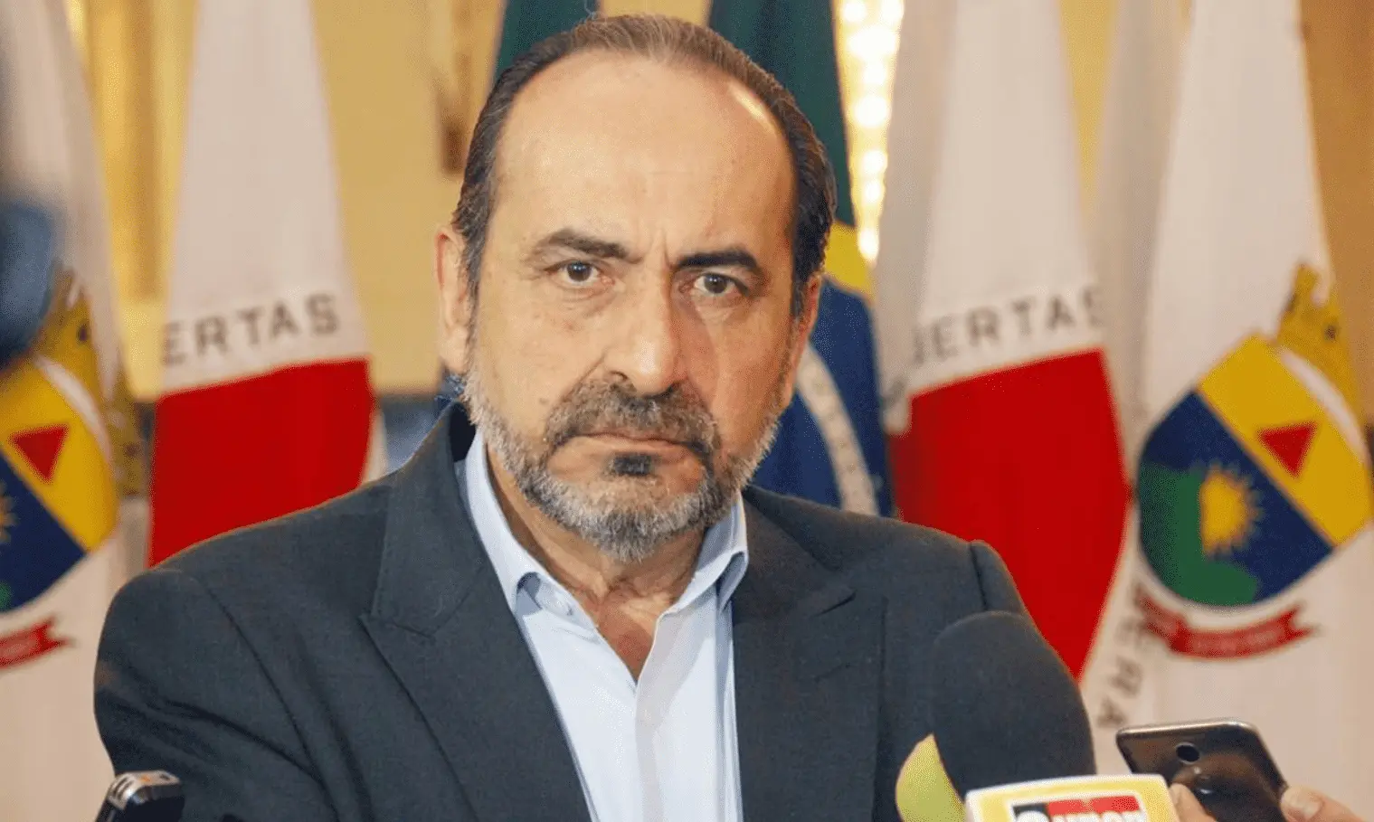 Foto mostra Alexandre Kalil de frente para câmera, usando terno. Ao fundo se vê bandeiras de MG. 