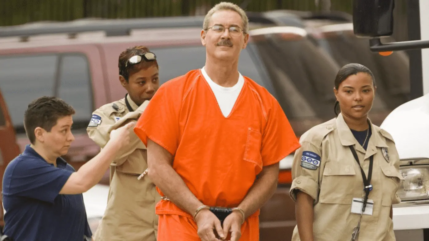 Ex-milionário sendo preso, vestindo uma roupa laranja, escoltado por policiais. 