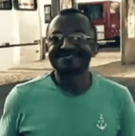 Foto mostra homem negro, de camiseta verde usando óculos e sorrindo. 