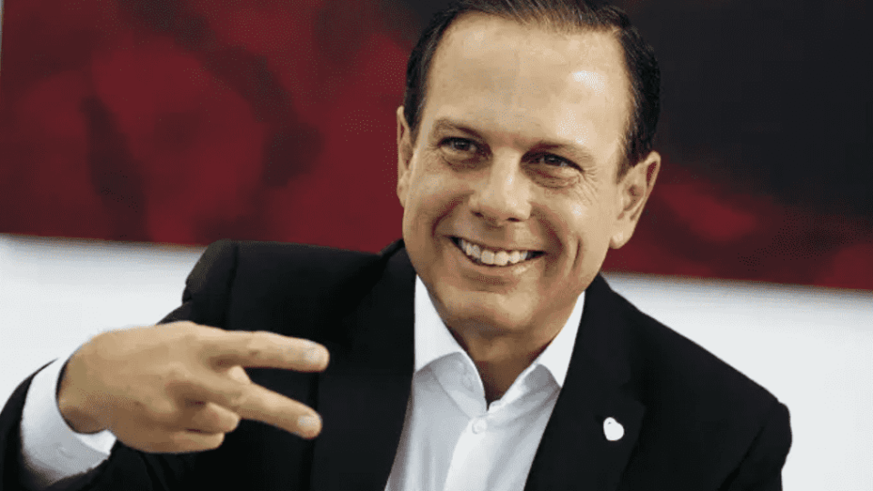 Foto mostra João Dória, do tronco para cima, fazendo um sinal com os dedos e sorrindo.