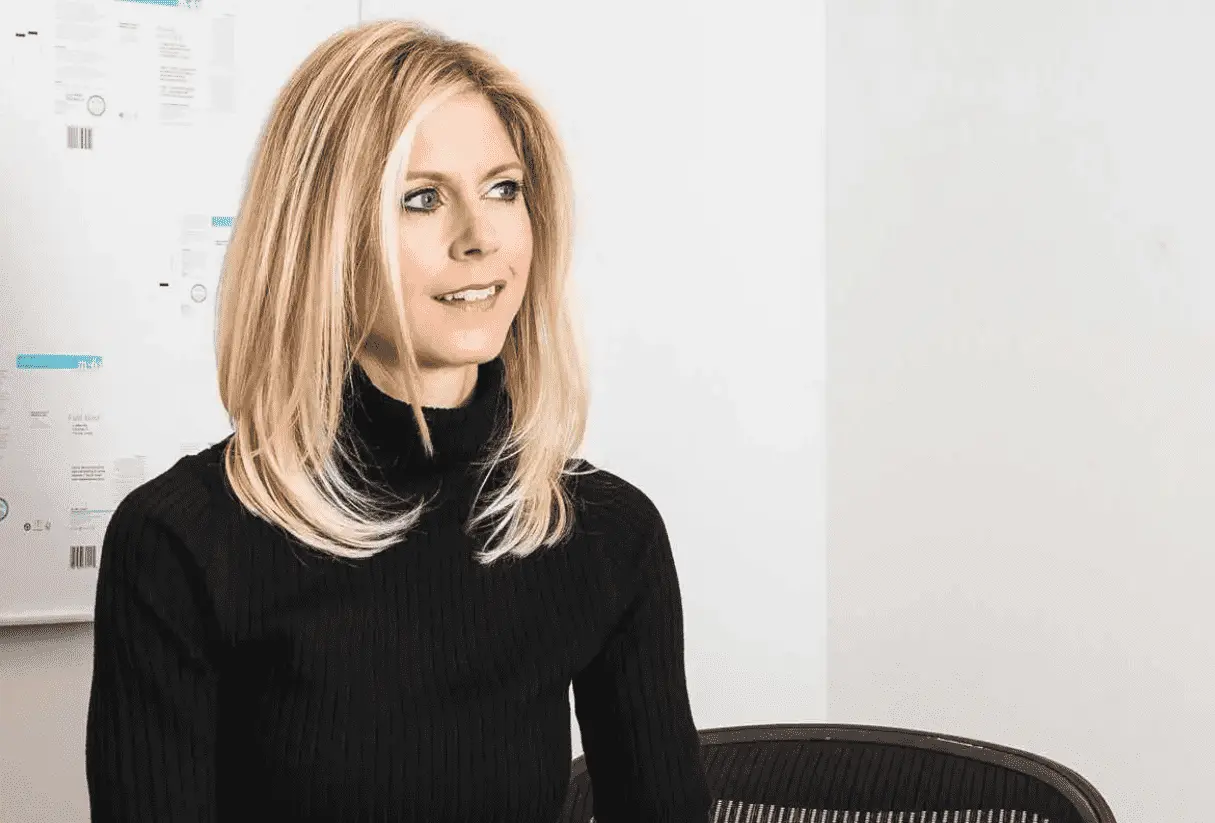 Foto mostra uma mulher adulta branca, de cabelos loiros, usando uma blusa de manga comprida preta.