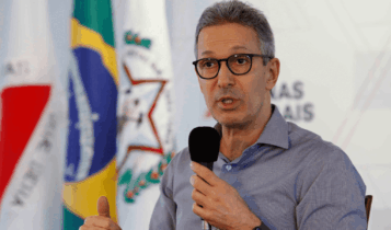 Foto mostra Romeu Zema, do tronco para cima, falando ao microfone. Ao fundo bandeiras do Brasil e de MG.