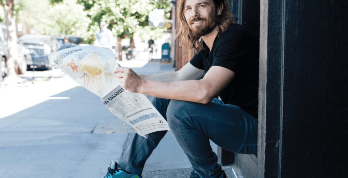 Foto mostra Dan Price sentado em um degrau, de lado, com um jornal nas mãos.