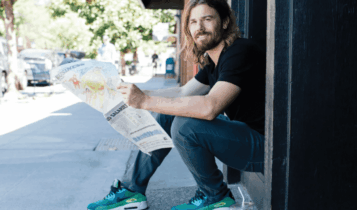 Foto mostra Dan Price sentado em um degrau, de lado, com um jornal nas mãos.