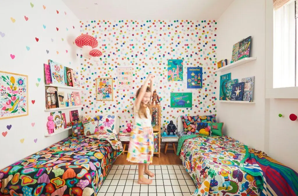crianças brincam no centro de seu quarto colorido entre duas camas e em frente a um papel de parede de confete