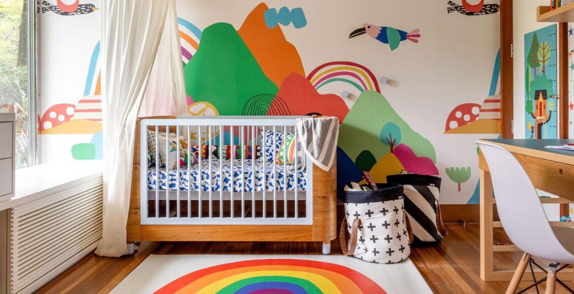 berço de bebê com roupa de cama colorida em frente a um papel de parede colorido de montanhas e passados.