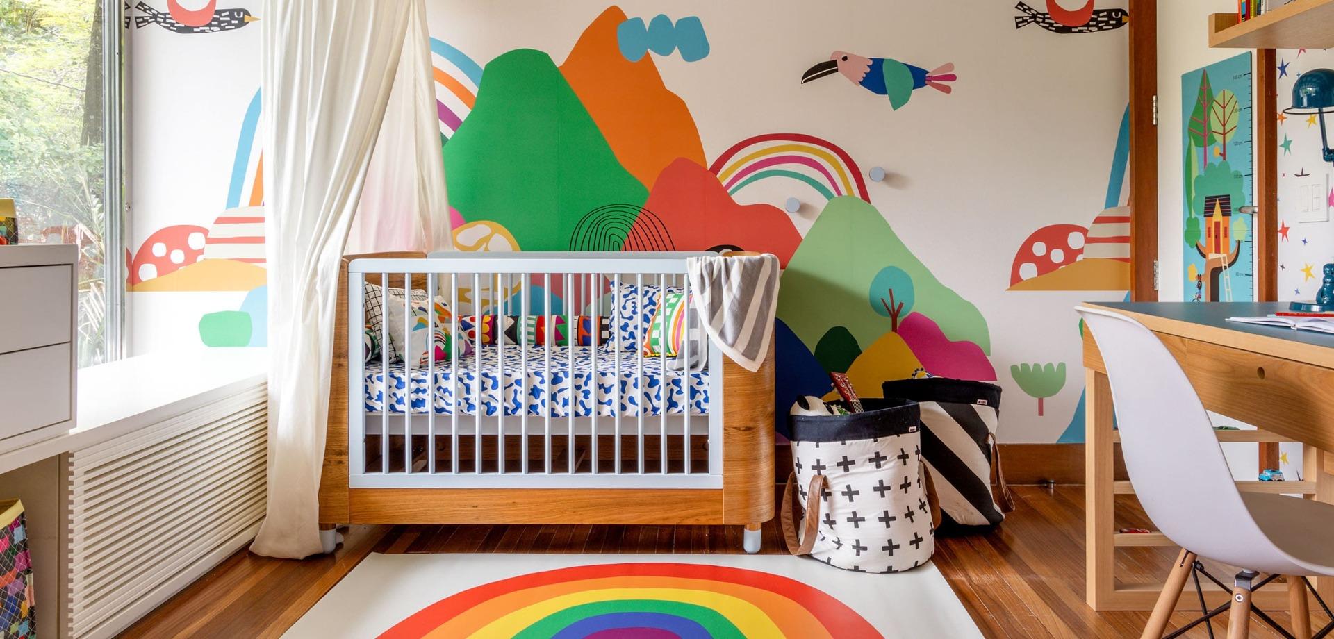 berço de bebê com roupa de cama colorida em frente a um papel de parede colorido de montanhas e passados.