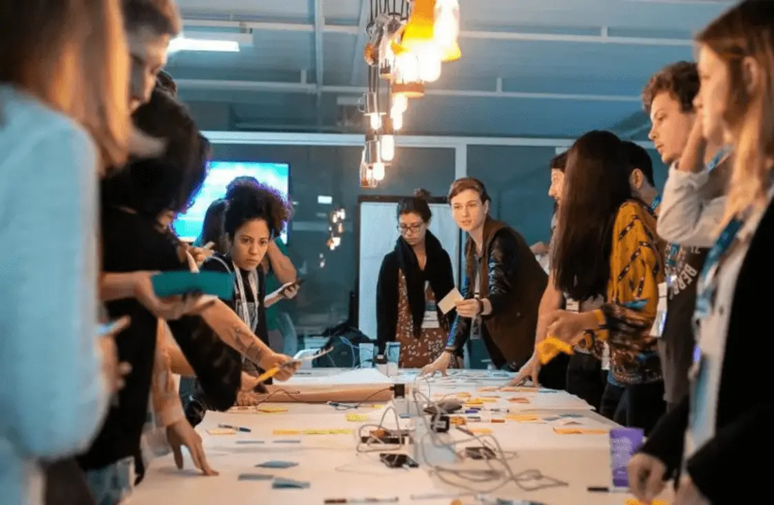 Foto mostra uma mesa com várias pessoas em volta em um ambiente empresarial. 