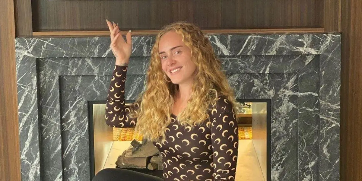 Adele agachada sorrindo com a mão direita levantada para o alto