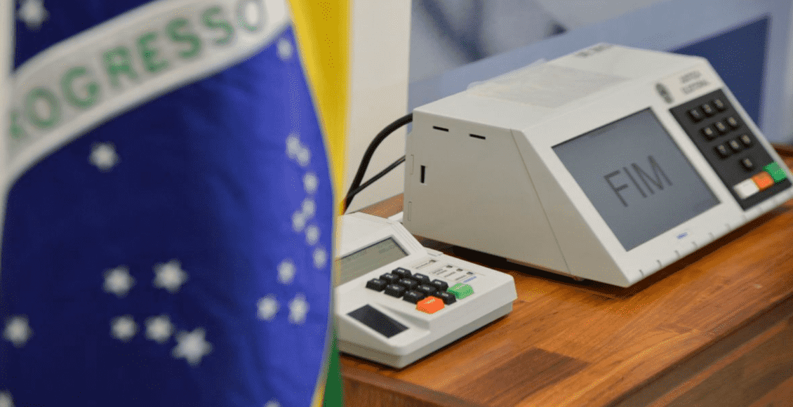 Foto mostra parte da bandeira do Brasil e uma urna eletrônica atrás.