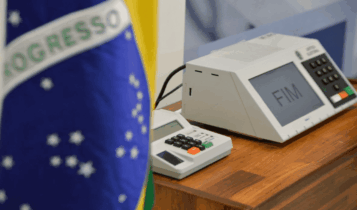 Foto mostra parte da bandeira do Brasil e uma urna eletrônica atrás.