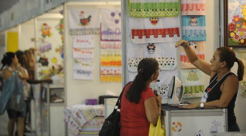 Foto mostra stands em uma feira de artesanato. Uma vendedora e uma cliente.