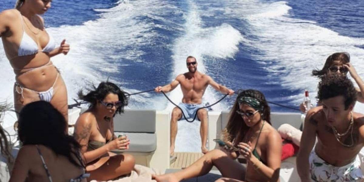 Anitta em um barco olhando para o celular. Ela está rodeada por amigos
