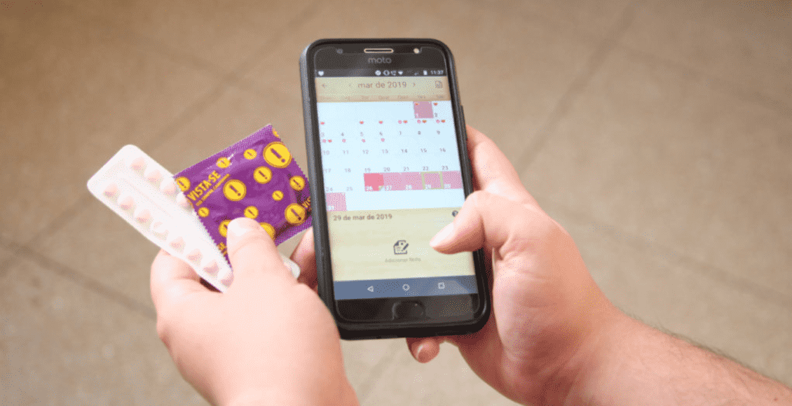Foto mostra uma mão segurando um celular, uma camisinha e uma cartela de anticoncepcional.