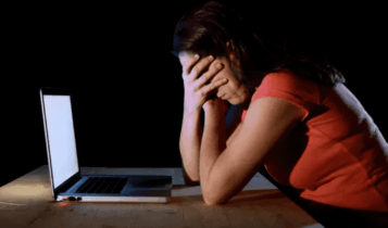 Foto mostra notebook na mesa e uma frente apoiada na frente com as mãos no rosto.