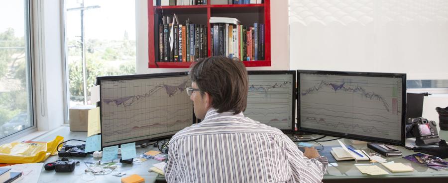 Investidor acompanhando três telas de computadores com gráficos de cotação