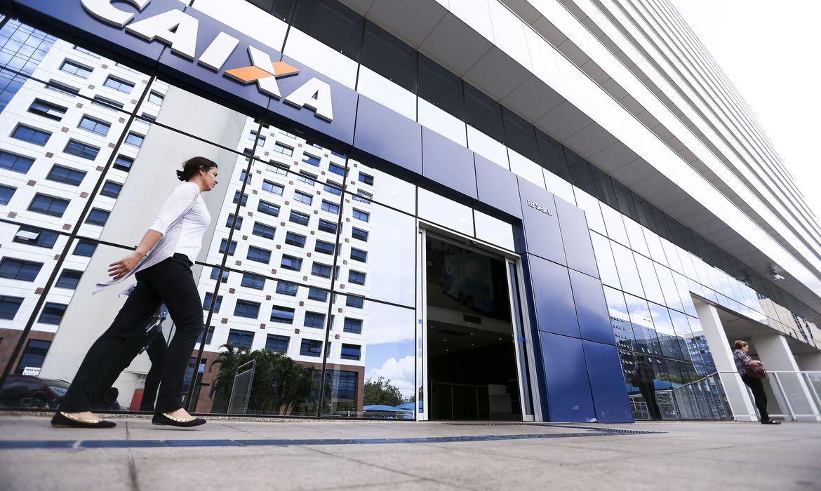 Fachada de uma agência caixa, com uma mulher de calça preta e camisa social branca andando ao lado esquerdo da imagem.