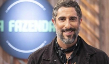 Marcos Mion posado sorrindo olhando para frente com letreiro de A Fazenda atrás