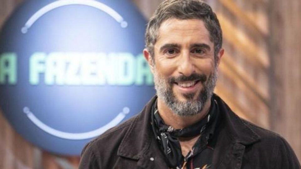 Marcos Mion posado sorrindo olhando para frente com letreiro de A Fazenda atrás