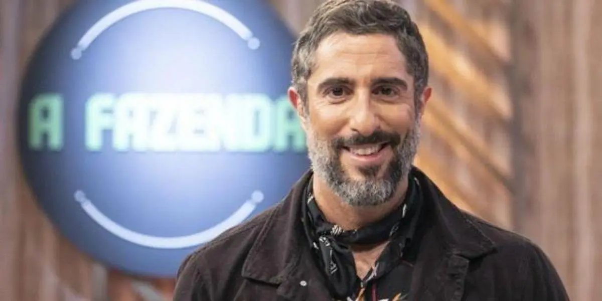 Marcos Mion posado sorrindo olhando para frente com letreiro de A Fazenda atrás