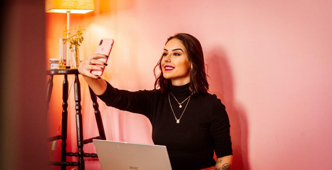 Imagem de mulher com notebook sobre o colo, segurando um celular com a mão direita e tirando uma selfie.
