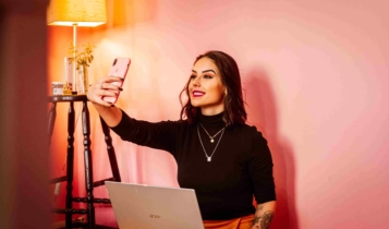 Filtros no Instagram: guia completo para criar o seu Imagem de mulher com notebook sobre o colo, segurando um celular com a mão direita e tirando uma selfie.