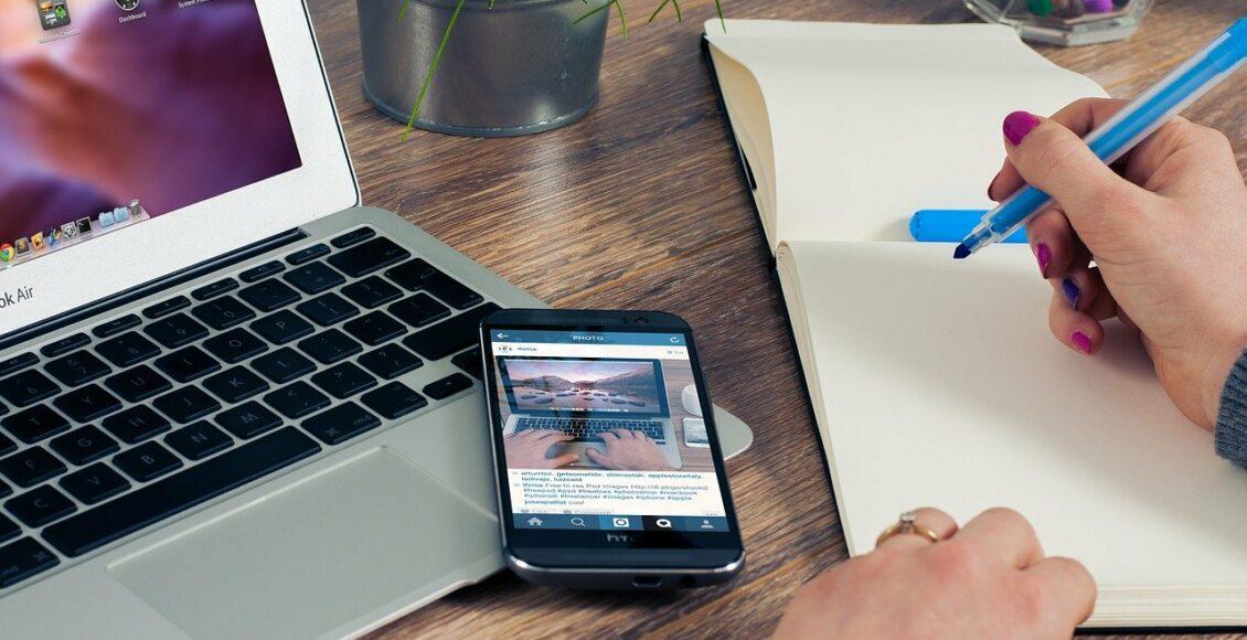 Como criar um bom portfólio online? Imagem mostra mulher anotando em caderno ao lato de celular e notebook