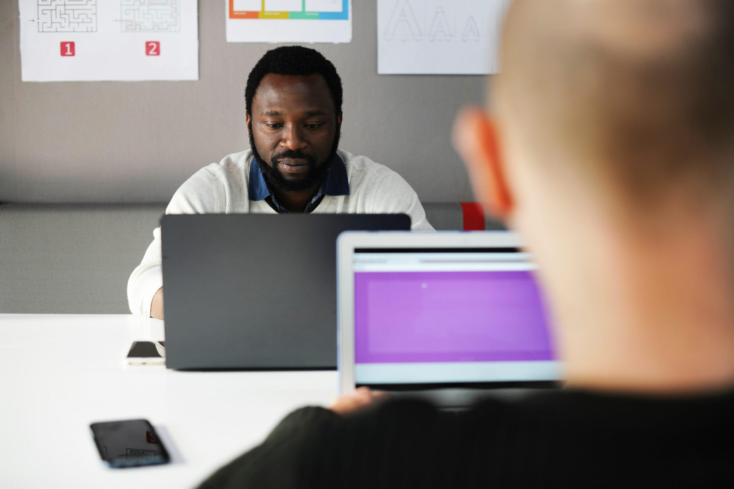 Dois homens sentados de frente para o computador sobre uma mesa, um em frente ao outro. Um está de costas para a foto e um de frente.
