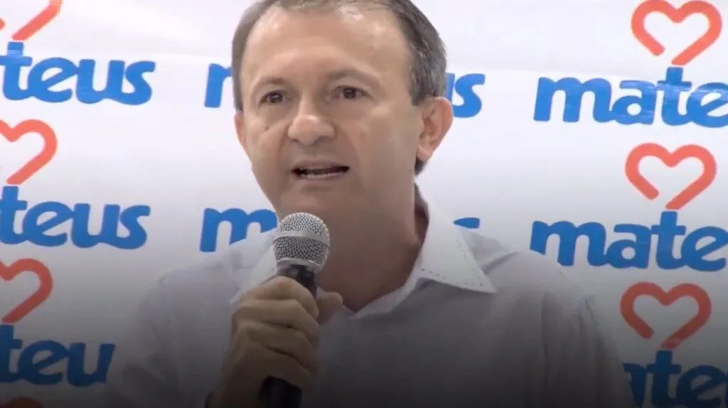 Ilson Mateus Ilson Mateus com microfone em uma das mãos e na frente de um painel com o logotipo do Grupo Mateus