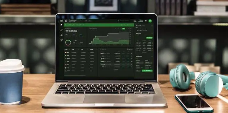 notebook com tela de trade em exchange de bitcoin, ao lado de um copo de café, iphone e phone de ouvido