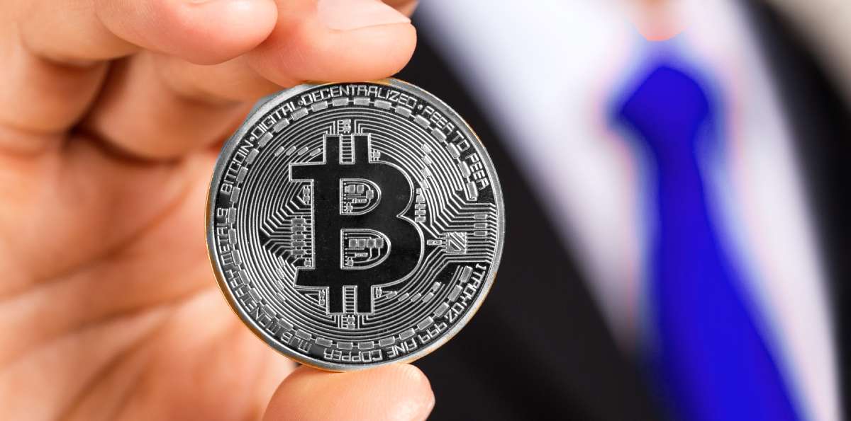 mão de pessoa usando terno e gravata azul segurando moeda com logotipo bitcoin