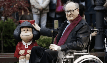 Foto mostra Quino ao lado de uma escultura da Mafalda