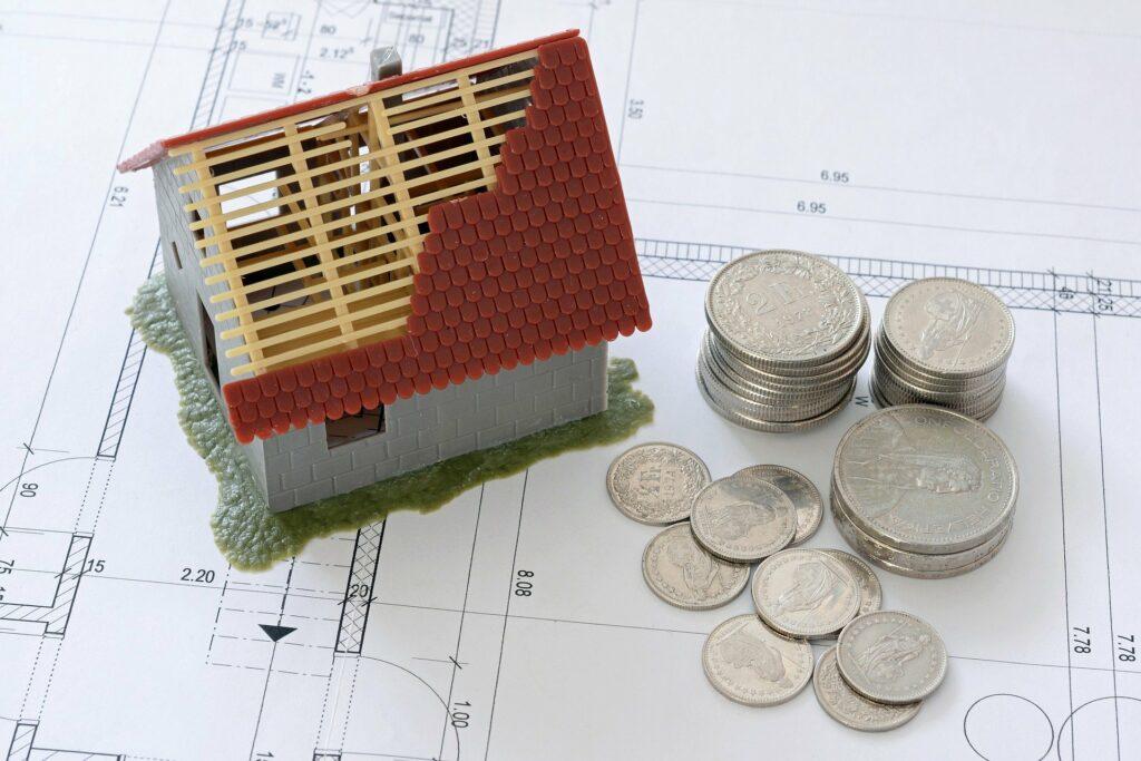 Miniatura de casa com parte do telhado sem cobertura, com moedas ao lado e sobre planilhas de dados