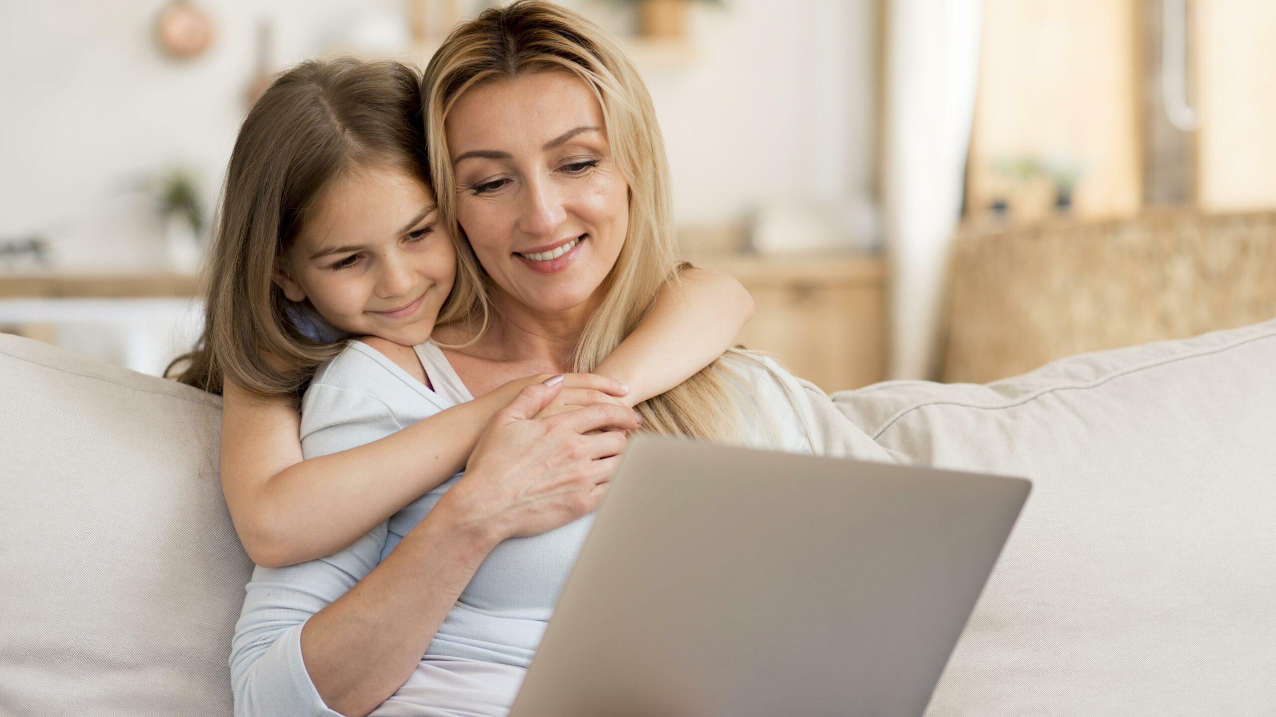Mulher usando notebook enquanto filha a abraça. Dependentes do MEI