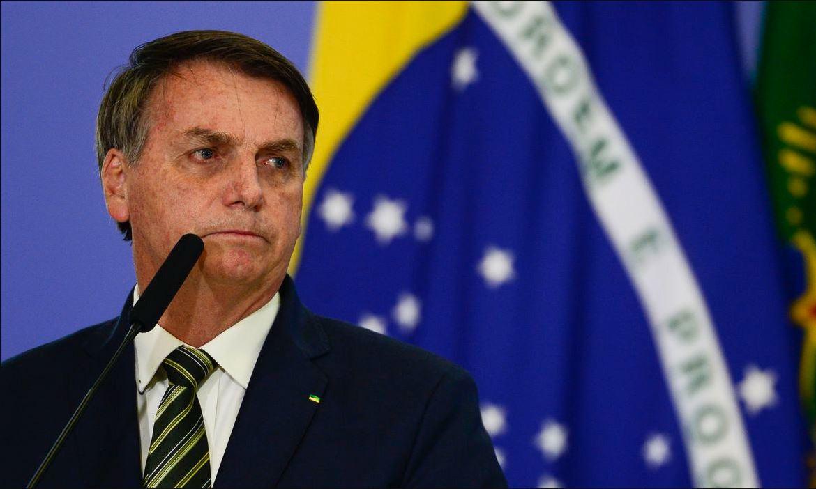 Foto mostra presidente Jair Bolsonaro alinhado a esquerda do enquadramento, vestido de terno, diante de um microfone e com olhar direcionado a direita (sua esquerda). Bandeira do Brasil se encontra ao fundo.