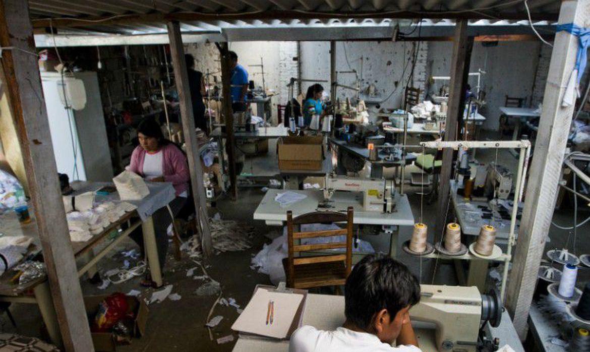 Foto mostra pessoas em situação de trabalho precárias em frente à máquinas de costura.