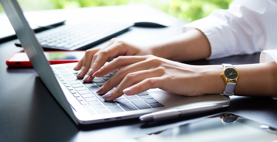 Foto mostra mãos de mulher branca digitando em teclado de notebook sobre a mesa.