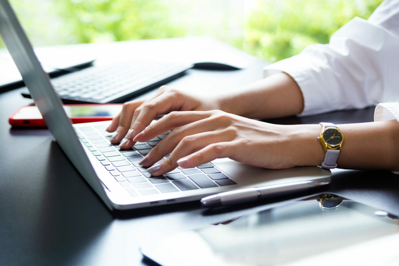 Foto mostra mãos de mulher branca digitando em teclado de notebook sobre a mesa.
