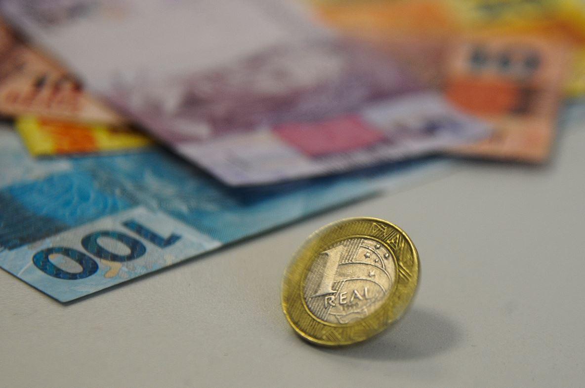 Foto mostra moeda de 1 real ao centro e notas de 100, 10 e 5 reais ao fundo.